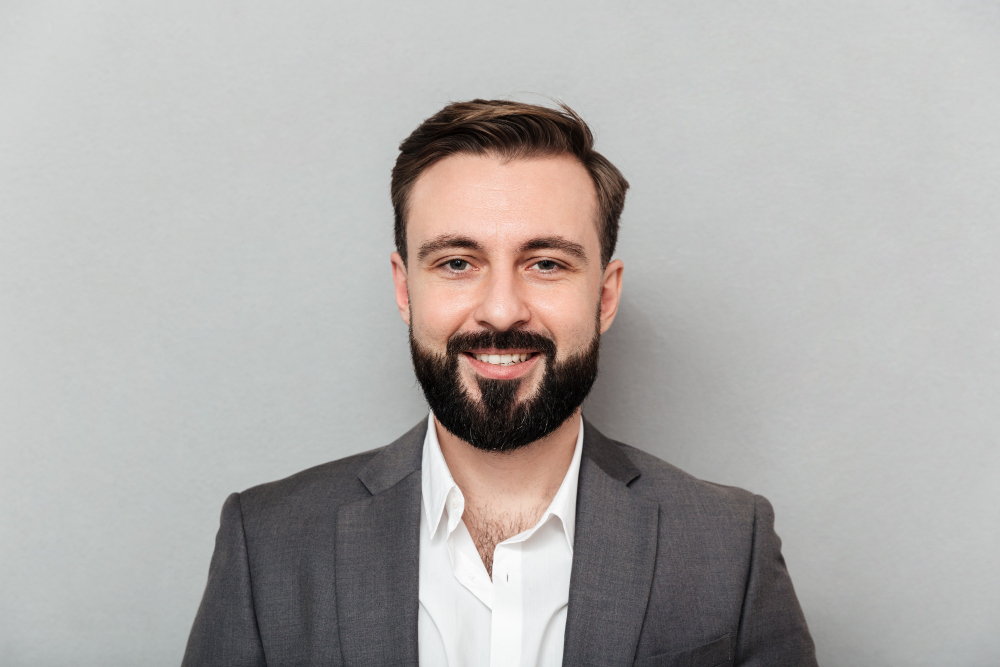 close-up-portrait-young-bearded-man-white-shirt-jacket-posing-camera-with-broad-smile-isolated-gray (1)